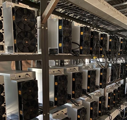 Rows of Antminer S19 units on steel shelving in a mining room, seen down the aisle with their power supplies sitting on top of each machine.