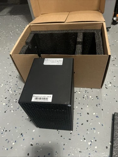A Goldshell Mini DOGE III miner on a speckled floor beside its opened carton and foam insert, a small matt-black box with a perforated intake panel.