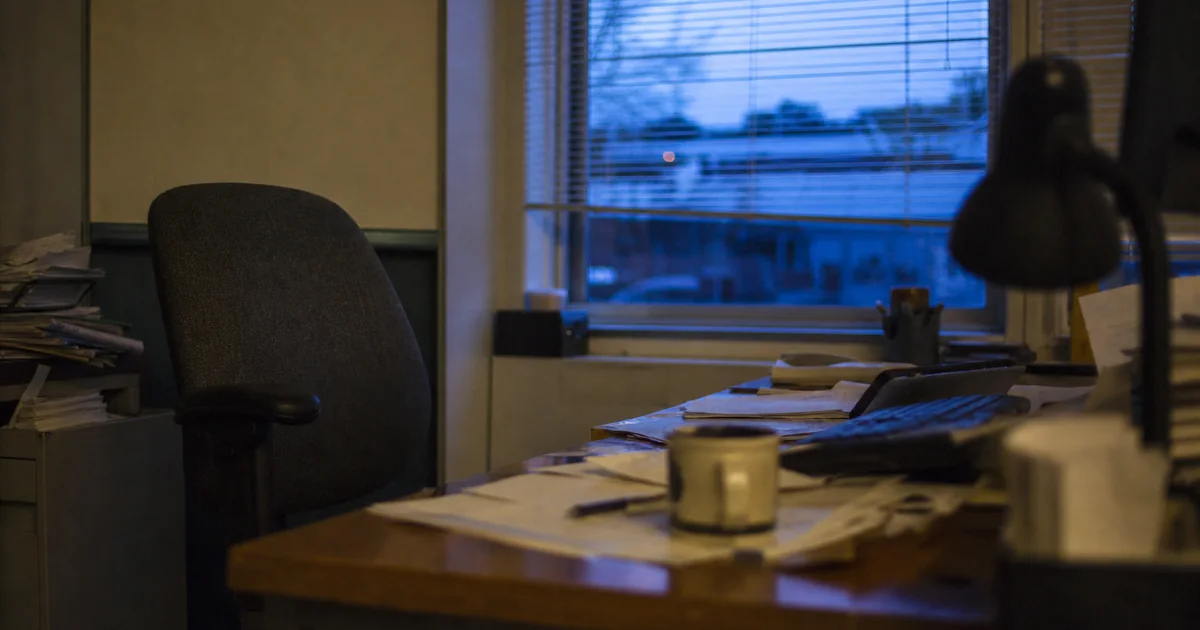 An empty office desk at dusk, chair pushed back, scattered paperwork and a cold mug of coffee.