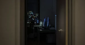A dark office seen through a doorway at night, a desk and its monitors just visible in the gloom.