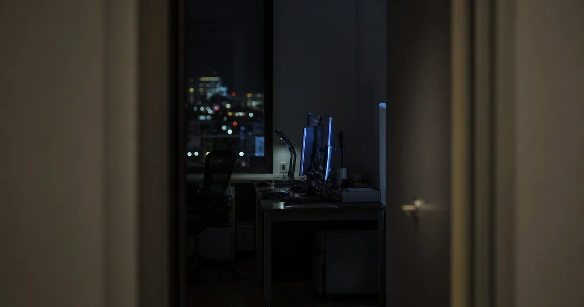 A dark office seen through a doorway at night, a desk and its monitors just visible in the gloom.