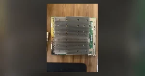 A used ASIC hashboard lying on a wooden desk, its aluminium heatsink block corroded and flux-stained at one corner, with test figures written on the green board edge in marker.