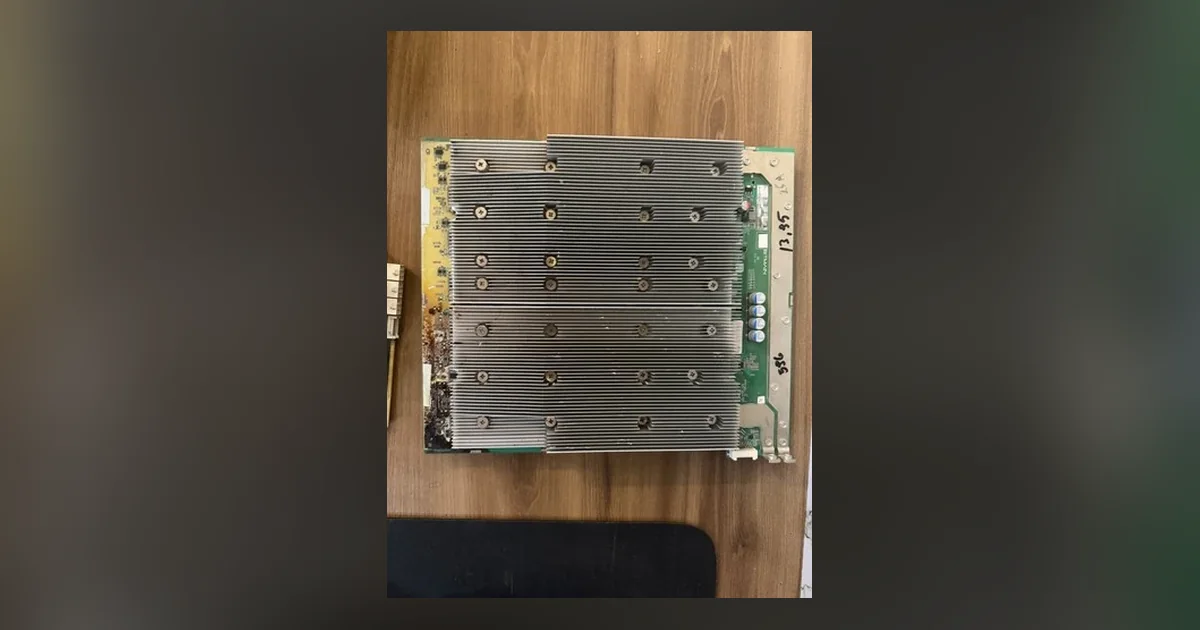 A used ASIC hashboard lying on a wooden desk, its aluminium heatsink block corroded and flux-stained at one corner, with test figures written on the green board edge in marker.