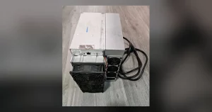 A refurbished Bitmain Antminer S19-series ASIC miner lying on a grey wood-effect floor, its control panel and Ethernet port facing the camera and its black power lead coiled alongside it.