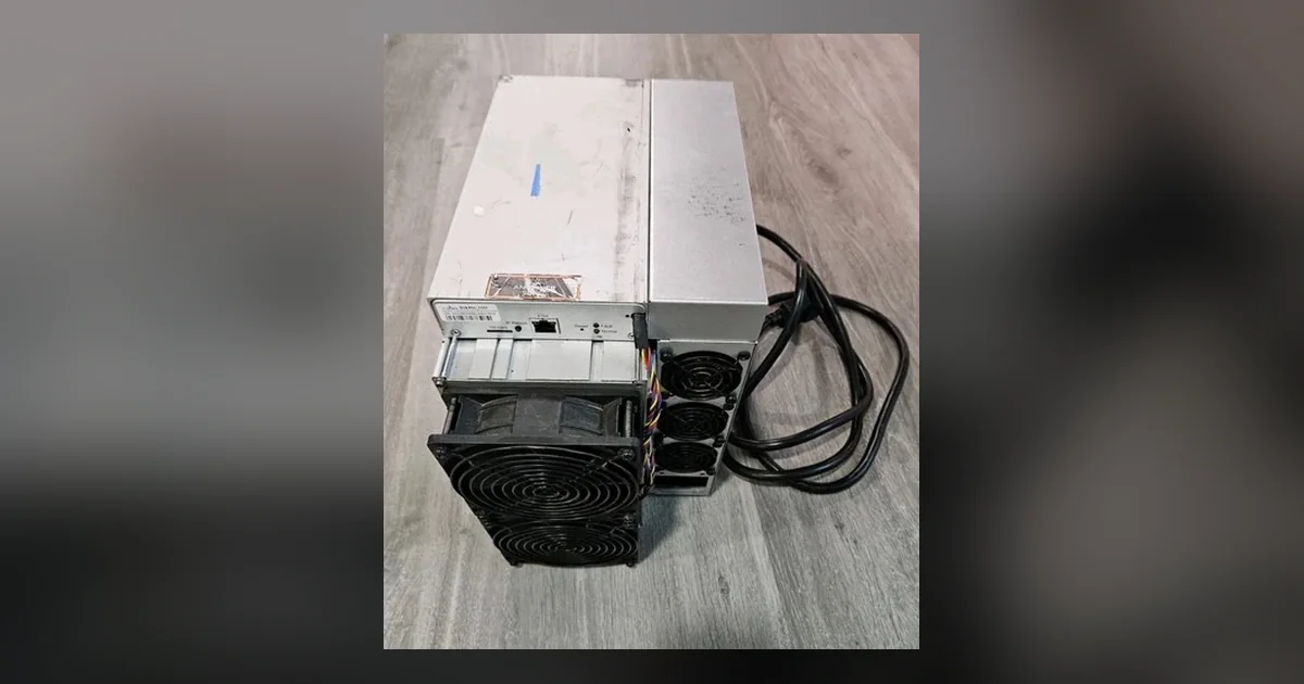 A refurbished Bitmain Antminer S19-series ASIC miner lying on a grey wood-effect floor, its control panel and Ethernet port facing the camera and its black power lead coiled alongside it.