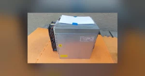 A Bitmain Antminer L9 Scrypt miner standing on an orange workshop mat, its ventilated steel case and end plate facing the camera.