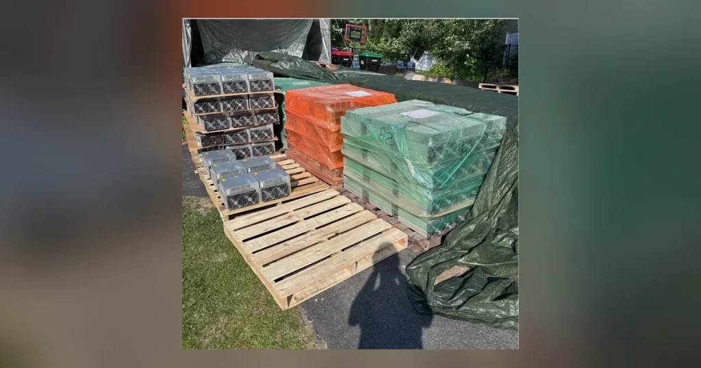 Pallets of ASIC miners shrink-wrapped in orange and green film, stacked on wooden pallets outdoors and ready for freight.