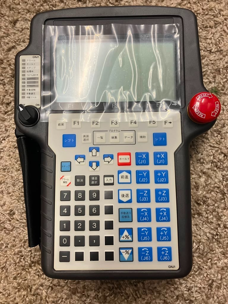 A FANUC teach pendant in its protective wrap, keypad and emergency-stop button visible.