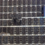 Used Antminer S19 units stacked in rows against a warehouse wall, fan grilles facing out, an old pedestal fan leaning on the stack.