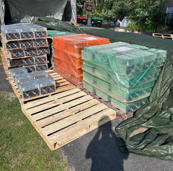Pallets of used Antminer S19 miners outdoors, bare units stacked in rows next to two shrink-wrapped pallets in orange and green plastic.