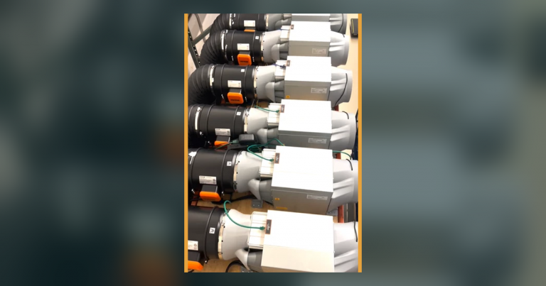 Our own photograph of six used Antminer S19k Pro miners racked in a row with flexible exhaust ducting fitted to each and their power supplies mounted alongside, the arrangement a multi-machine used fleet actually takes up.