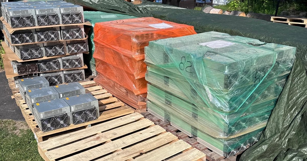 Used Bitmain Antminer S19 miners on wooden pallets, some stacked bare and some shrink-wrapped in orange and green with paperwork taped to the load, ready for freight.