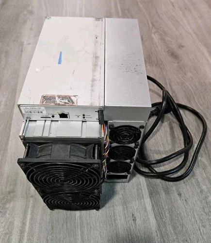 A used Bitmain Antminer S19 standing upright on a grey wood-effect floor, seen from above: two stacked black intake fans and the control board on its front face, the silver power supply attached at the rear with its mains lead coiled beside it, and a worn label on the top of the case.