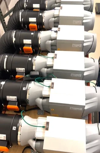 A row of used Antminer S19k Pro machines shelved end-on, each showing a black round fan housing beside a pale power supply box, with orange clips and green leads running between them.