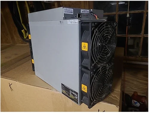 A used Bitmain Antminer S19K Pro standing on a cardboard box in a wood-panelled room, its control board and wiring loom across the top and two stacked intake fans with yellow warning triangles facing right.