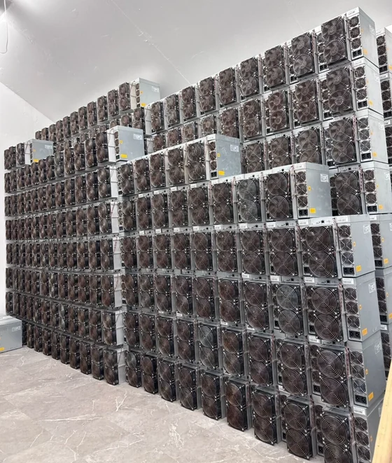 A wall of used Bitmain Antminer S19 machines stacked floor to ceiling across a tiled room, every unit turned so its fan face shows, with dust on the grilles and white stock labels on many of the cases.
