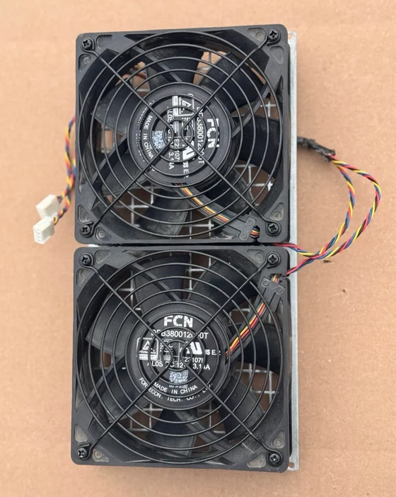 Two 120mm FCN case fans mounted one above the other on a shared black backplate with wire guards over both, their four-wire looms and white connectors trailing to one side, lying on brown cardboard.