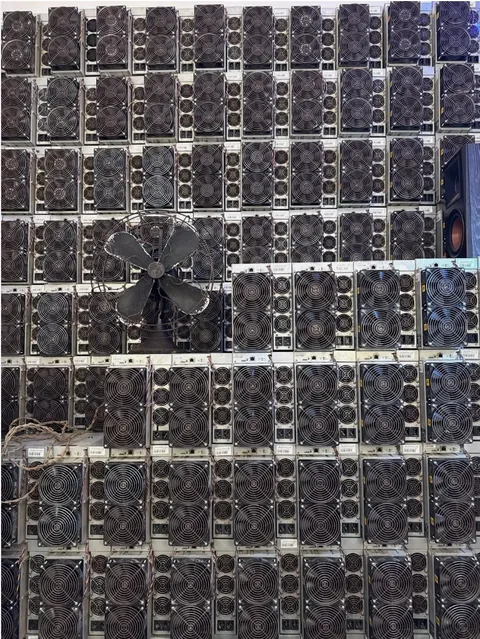 Not either machine in this comparison: a wall of refurbished Bitmain Antminer S19 units stacked in a storage room, fan faces out, with a free-standing floor fan pushing air across them.