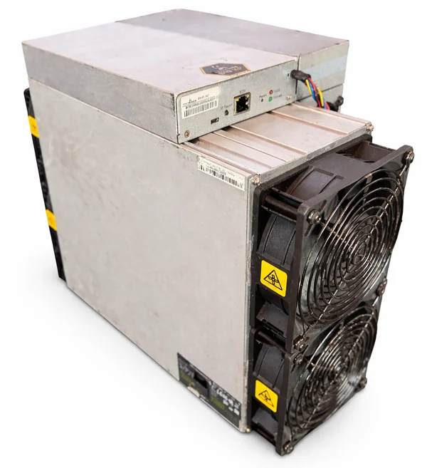 A used Bitmain Antminer S19k Pro standing upright on a pale floor, its silver power supply mounted along the top of the case and two large black intake fans filling the front face.