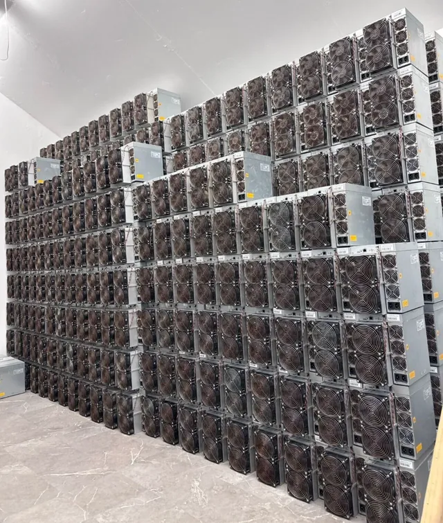 Used Bitmain Antminer S19 miners stacked several rows high against a warehouse wall, power supplies resting on top of each row.