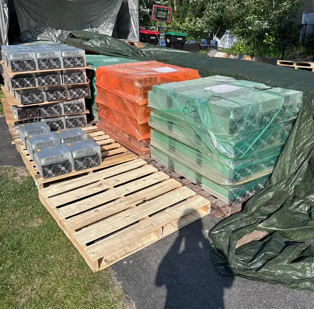 Used Bitmain Antminer S19 miners stacked and shrink-wrapped in orange and green on wooden pallets outside on grass.