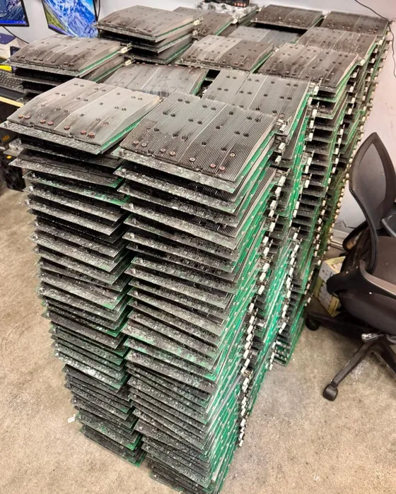 A tall stack of used Bitmain Antminer hashboards pulled from dead machines, standing on a workshop floor, heatsink faces dusty and green PCB edges showing.