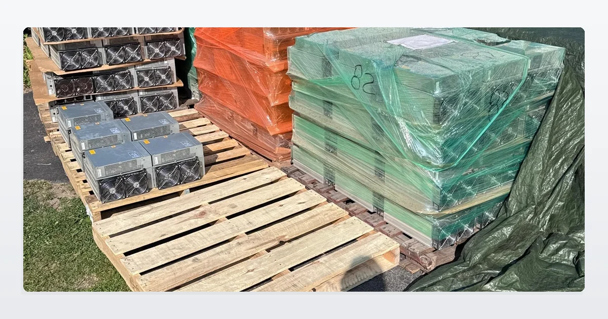 Used Bitmain Antminer S19 miners shrink-wrapped in orange and green on wooden pallets outdoors, with bare units stacked beside them and a pallet standing empty in front.