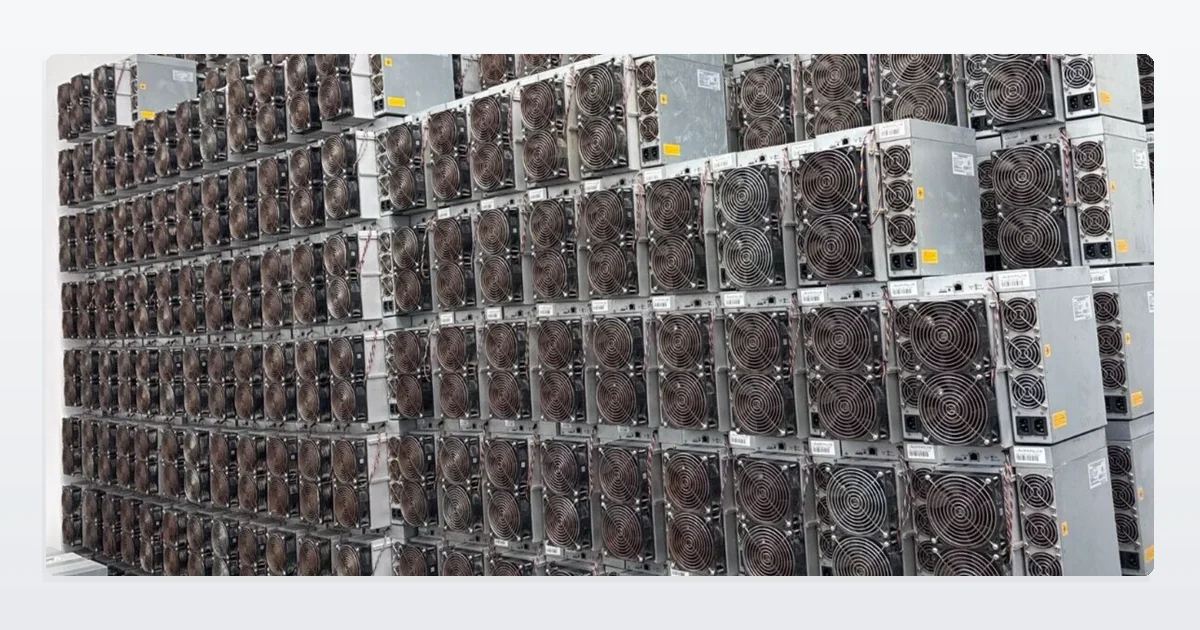 Used Bitmain Antminer S19 miners stacked six rows high against a warehouse wall on a concrete floor, power supplies resting on top of each row.