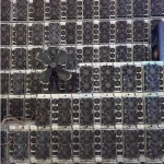 Refurbished Bitmain Antminer S19 110TH/s miners stacked in dense rows against a warehouse wall, fan grilles facing out, with an old pedestal fan standing against the stack.