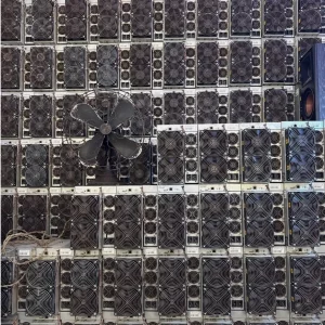 Refurbished Bitmain Antminer S19 110TH/s miners stacked in dense rows against a warehouse wall, fan grilles facing out, with an old pedestal fan standing against the stack.